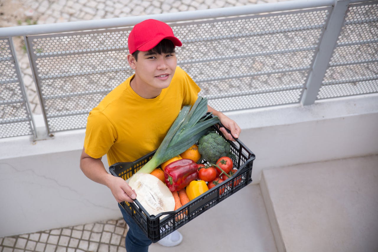 Young man holding a crate of fresh vegetables outdoors, wearing a red cap and yellow shirt.
